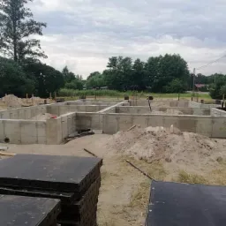 Wylany betonowy fundament domu jednorodzinnego z widocznymi zbrojeniami, sterta piasku i czarne szalunki na pierwszym planie, pochmurne niebo w tle.