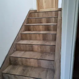 Schody obłożone płytkami imitującymi drewno, z ciemnymi listwami wykończeniowymi, prowadzące do drewnianych drzwi w białej futrynie.