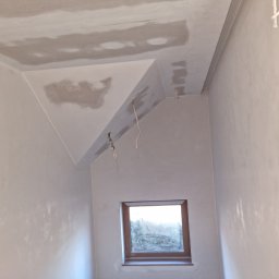 Darek sopel - Pokój ze skośnym sufitem w trakcie szpachlowania; widoczne niedoskonałości na ścianach i suficie, wystające kable elektryczne, okno z widokiem na zewnątrz.