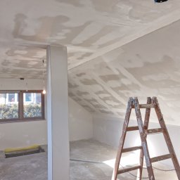 Darek sopel - Poddasze w trakcie prac wykończeniowych: ściany i sufit pokryte warstwą szpachli, drewniana drabina oparta o ścianę z surowej cegły, widoczne okno i kolumna.