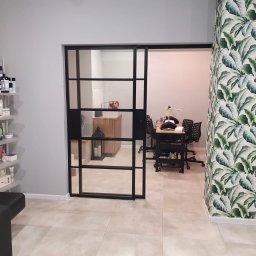 Darek sopel - Wnętrze salonu kosmetycznego z przeszklonymi przesuwnymi drzwiami w czarnej ramie, widoczne stanowisko do manicure z lampą, regał z kosmetykami Alfaparf i ściana w tapetę z motywem liści bananowca.