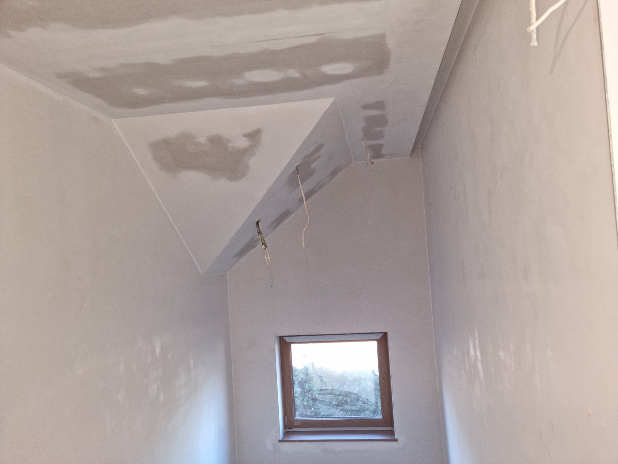 Pokój ze skośnym sufitem w trakcie szpachlowania; widoczne niedoskonałości na ścianach i suficie, wystające kable elektryczne, okno z widokiem na zewnątrz.