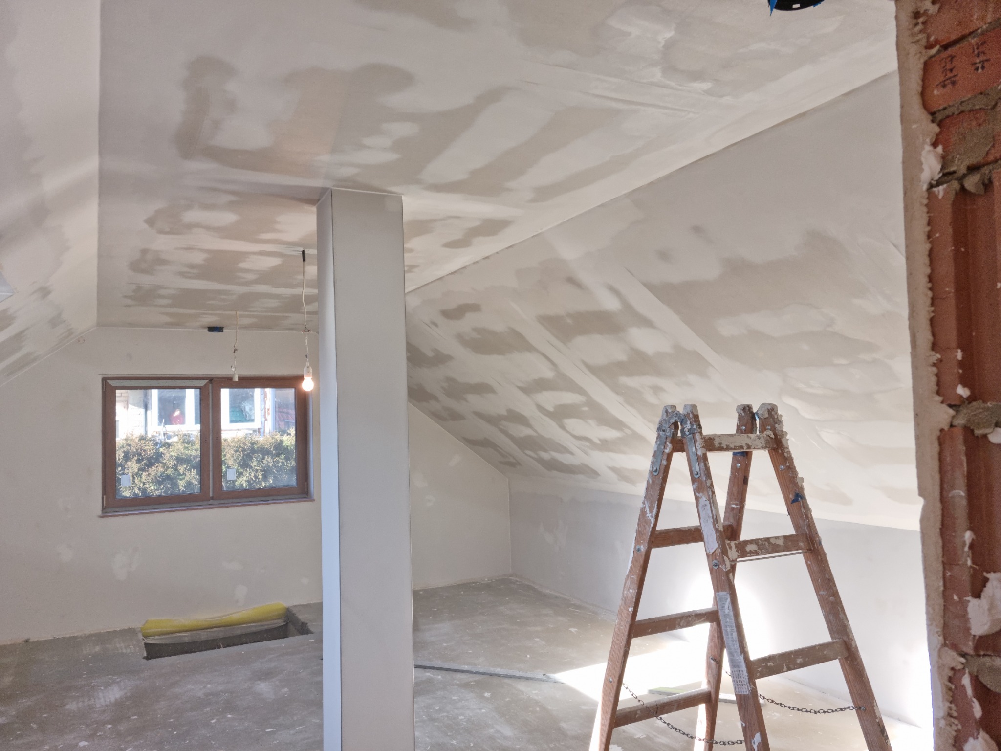 Poddasze w trakcie prac wykończeniowych: ściany i sufit pokryte warstwą szpachli, drewniana drabina oparta o ścianę z surowej cegły, widoczne okno i kolumna.