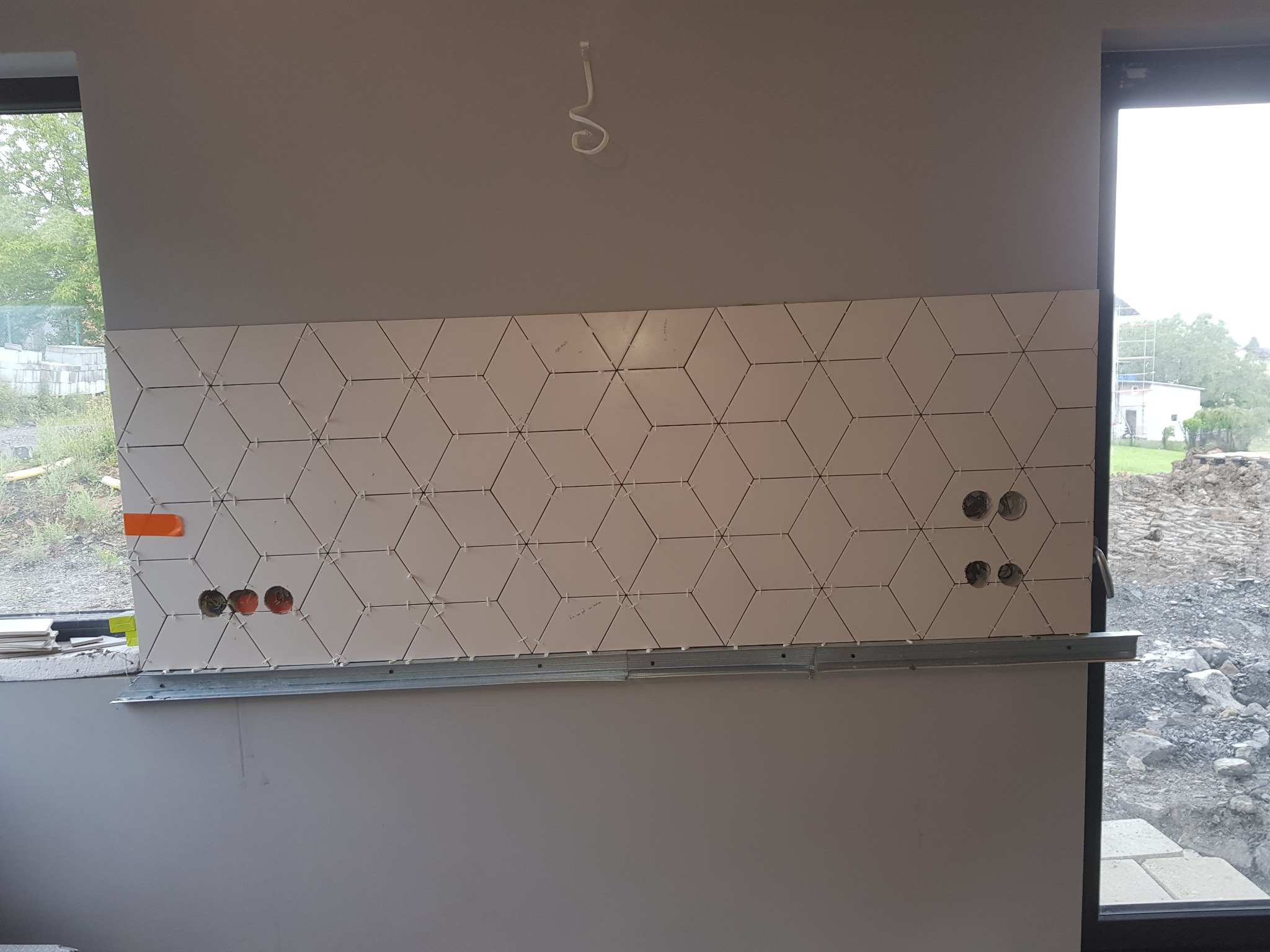 Montaż białych, sześciokątnych płytek z dystansami na ścianie z otworami na gniazdka, podtrzymywanych przez metalowy profil, widok przez okno na zewnątrz.