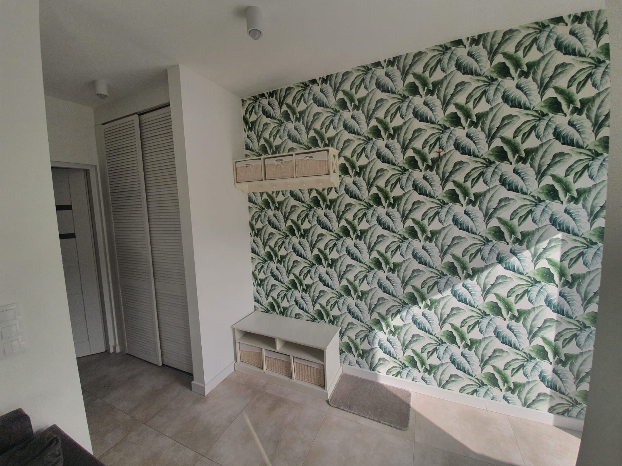 Nowoczesny przedpokój z tapetą w motywy liści, białą szafką z koszykami i płytkami podłogowymi w odcieniach beżu.