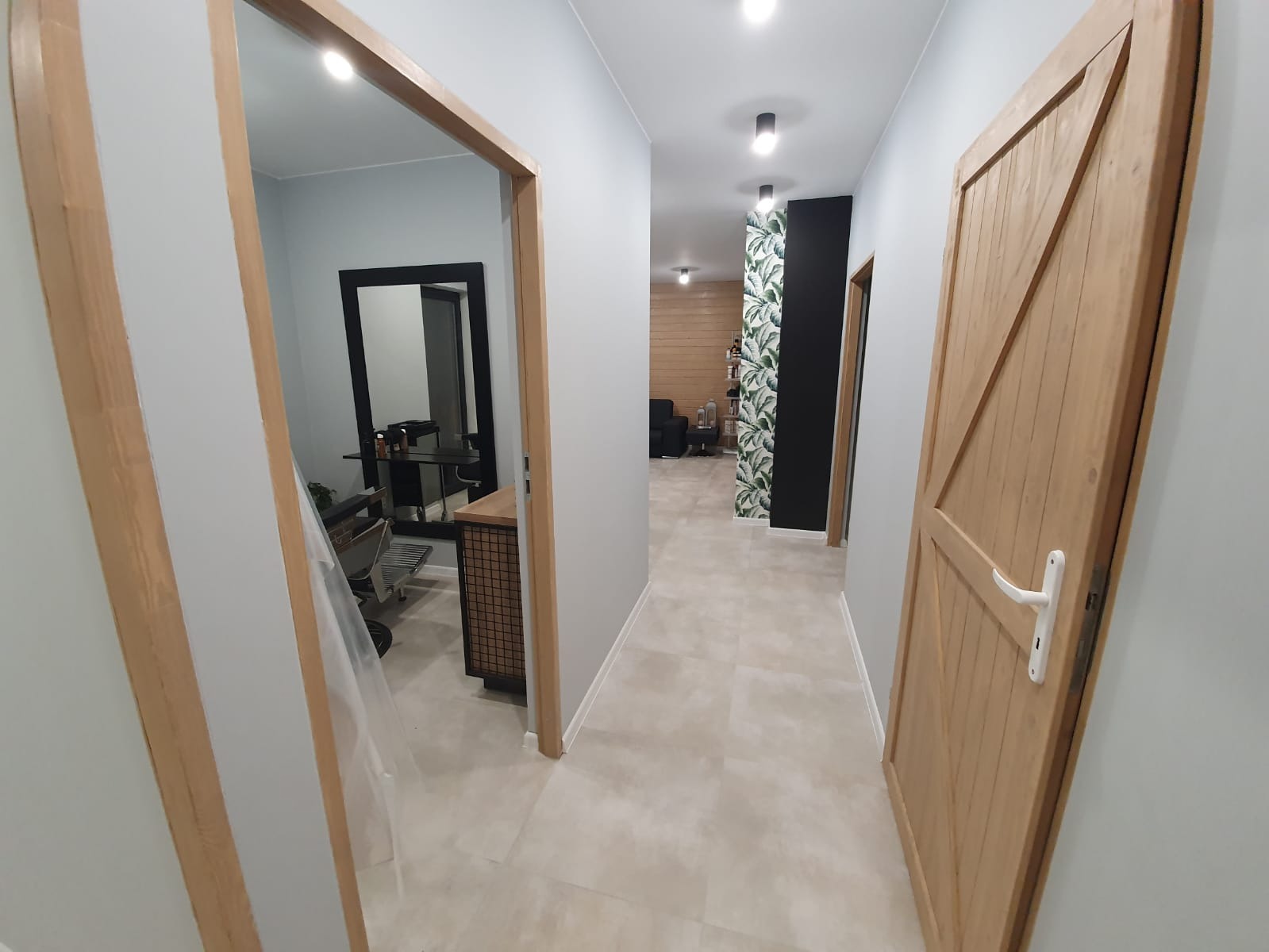 Wykończony korytarz z drewnianymi ościeżnicami, drzwiami w stylu rustykalnym i podłogą z jasnych płytek, widok na salon z drewnianą boazerią i ścianą z motywem roślinnym.