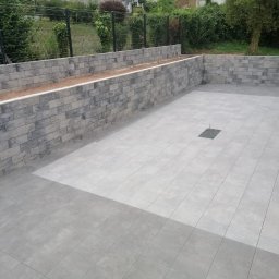 Ipo Sp. z o. o. - Wykończony taras z szarych płytek ceramicznych, obrzeżony murem z bloczków imitujących kamień, z centralnie umieszczoną kratką odpływową.