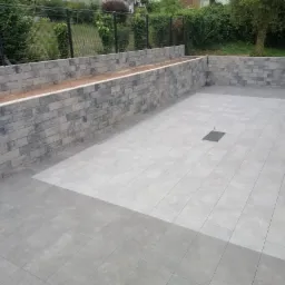 Wykończony taras z szarych płytek ceramicznych, obrzeżony murem z bloczków imitujących kamień, z centralnie umieszczoną kratką odpływową.
