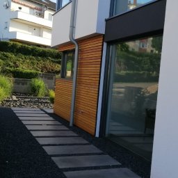 Ipo Sp. z o. o. - Nowoczesny dom z elewacją z drewnianych paneli i ścieżką z płyt na czarnym żwirze, widok z boku na tle zieleni.