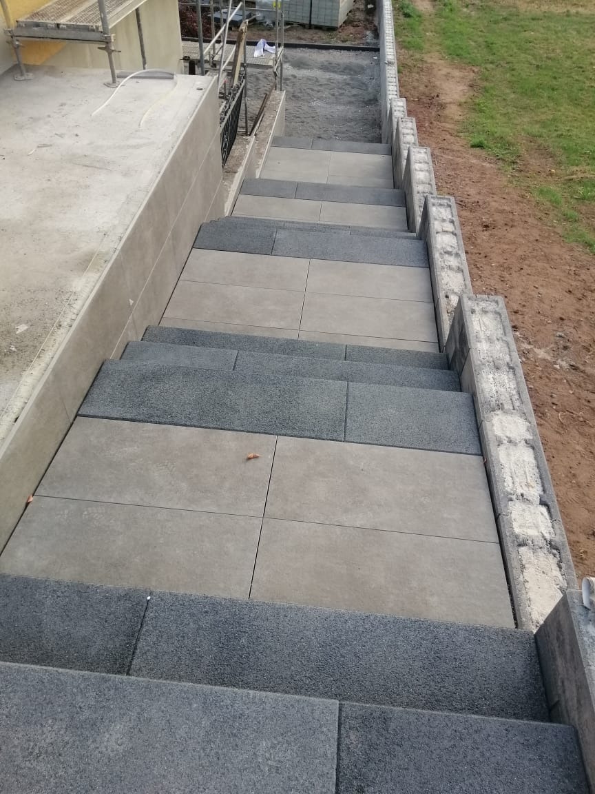 Schody zewnętrzne wyłożone kombinacją jasnoszarych płyt i ciemnoszarej kostki z fakturą, widok z góry na dół, z elementami rusztowania w tle.