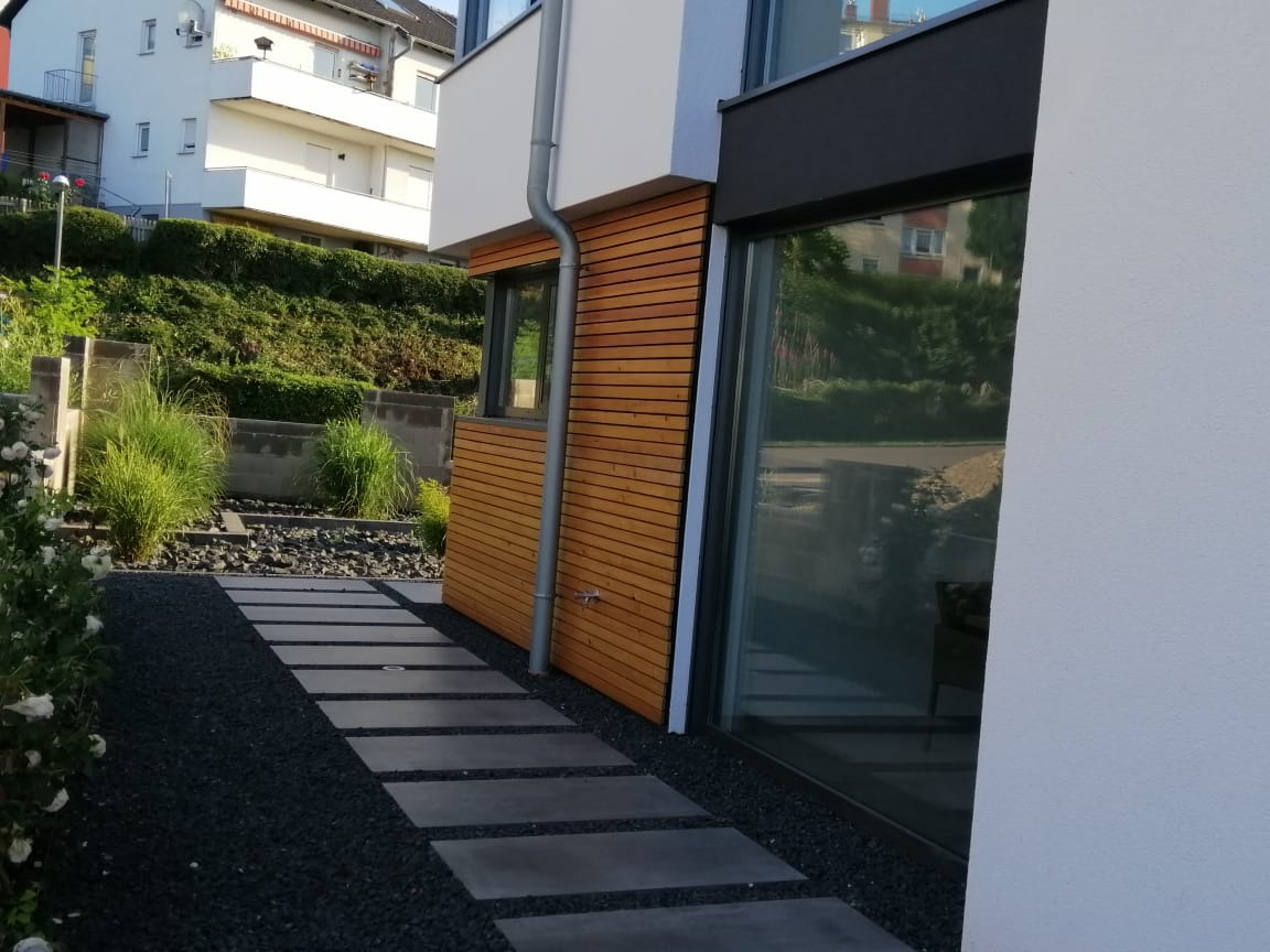 Nowoczesny dom z elewacją z drewnianych paneli i ścieżką z płyt na czarnym żwirze, widok z boku na tle zieleni.