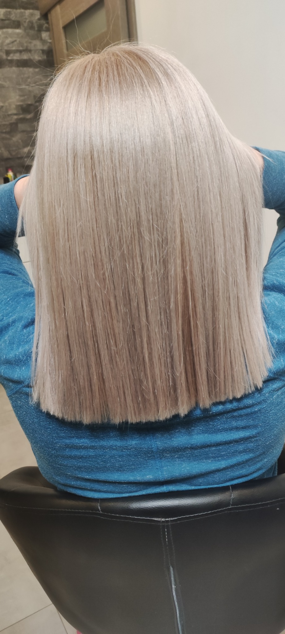 Proste, platynowe blond włosy średniej długości, widziane z tyłu, na osobie w niebieskiej bluzce siedzącej na czarnym krześle.