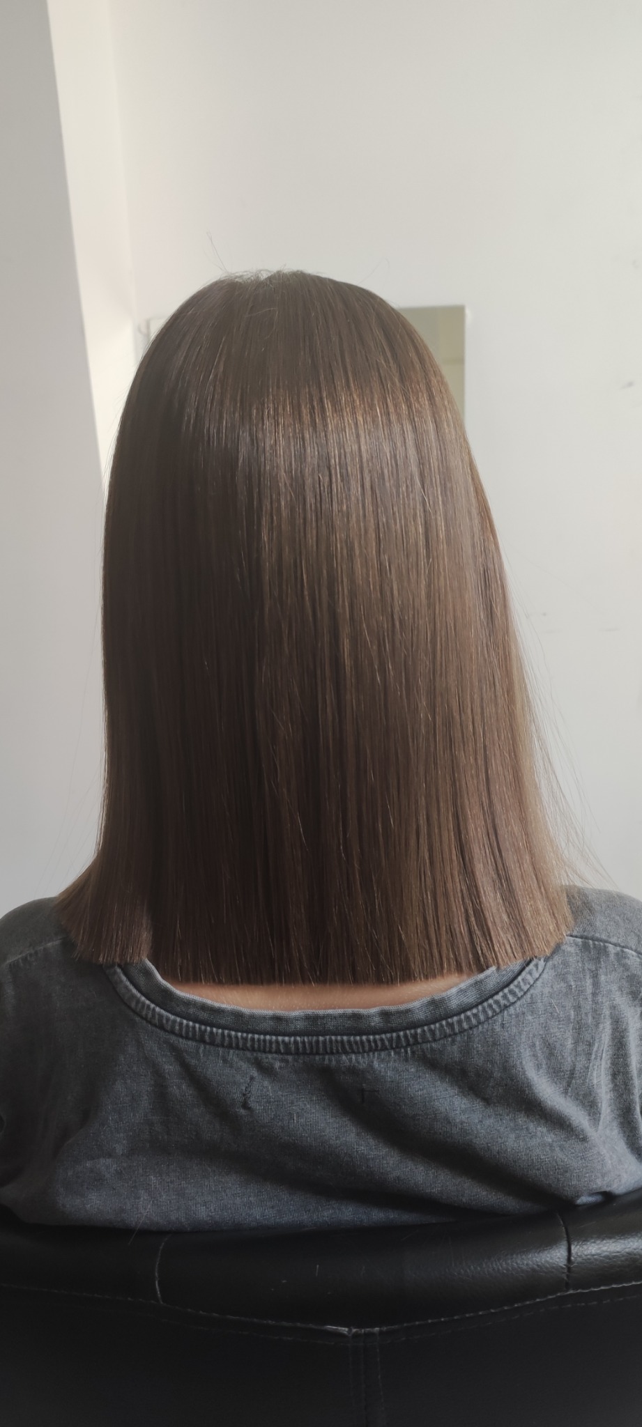 Tył głowy z brązowymi, prostymi włosami o długości do ramion, świeżo po strzyżeniu, widoczne lekkie refleksy, t-shirt w odcieniach szarości.