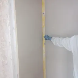 Sprawdzanie pionu świeżo nałożonej, jasnej ściany za pomocą poziomicy przez osobę w kombinezonie ochronnym i niebieskich rękawicach.