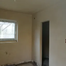 Wnętrze w trakcie prac wykończeniowych: ściany pokryte świeżym tynkiem, widoczne rury grzewcze na podłodze, okno z widokiem na zewnątrz, surowy stan wykończenia.