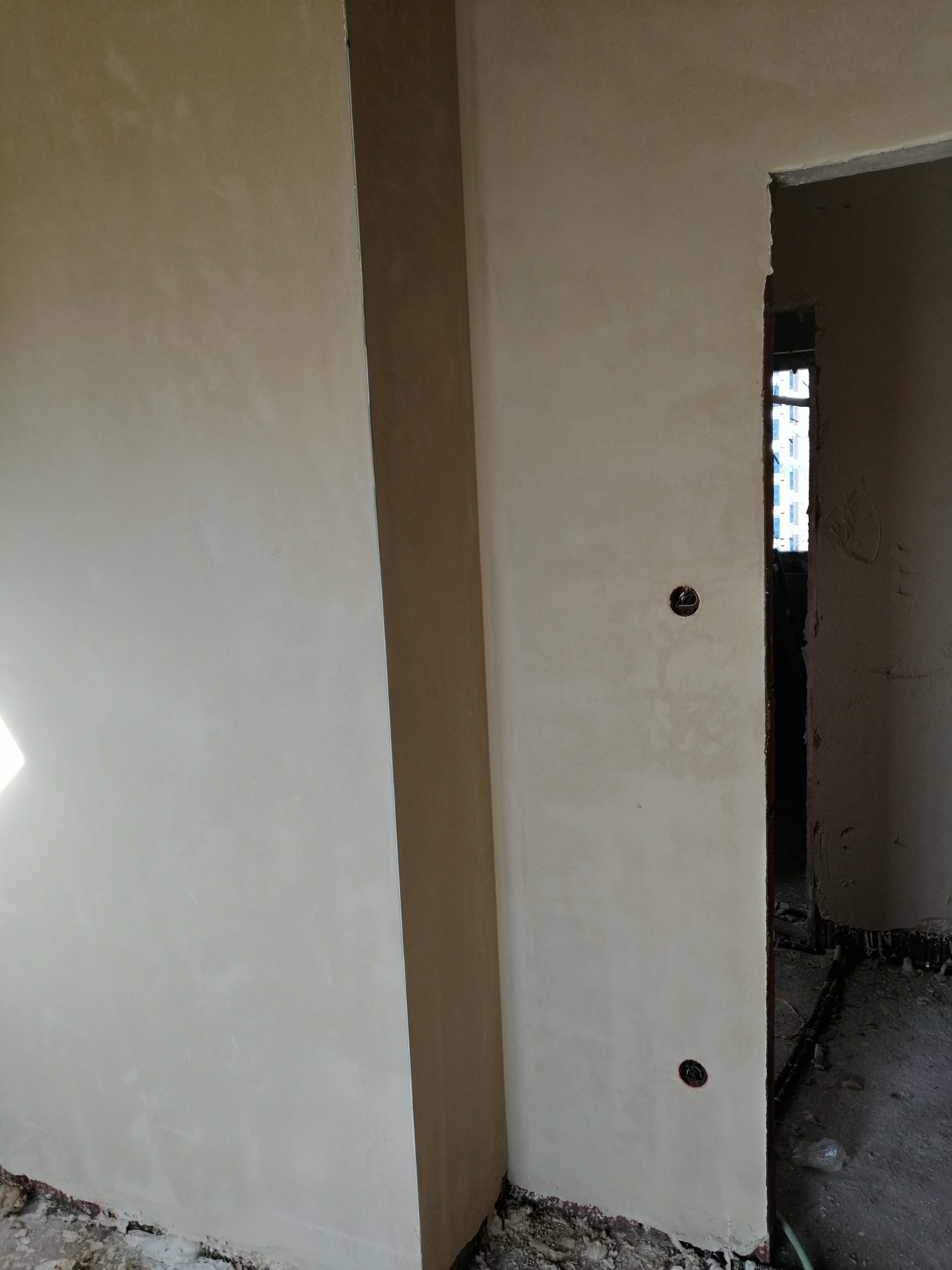 Ściana po nałożeniu warstwy wykończeniowej, widoczne gniazdka elektryczne i surowe wykończenie ościeżnicy, fragment podłogi z gruzem.