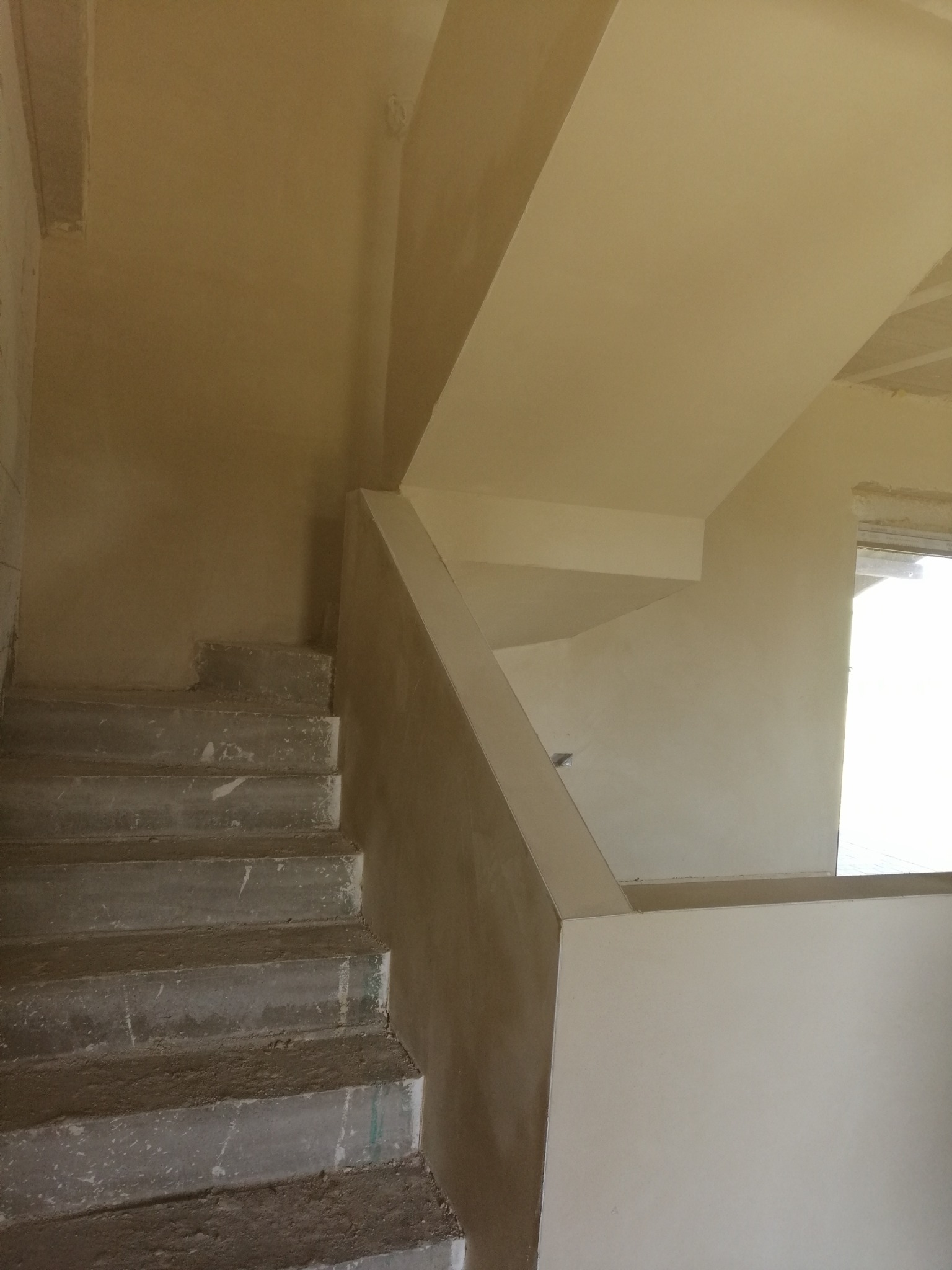 Surowe betonowe schody w trakcie remontu, ściany pokryte świeżym tynkiem w odcieniach beżu, widoczne niedoskonałości i brak wykończenia, naturalne światło wpadające przez okno.