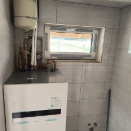 Eco Therm - Biała pompa ciepła Panasonic Aquarea z zbiornikiem wyrównawczym, zamontowana w pomieszczeniu z szarymi płytkami, widok z przodu, instalacja w trakcie wykańczania.