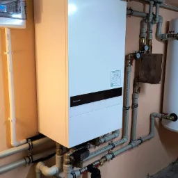 Biały piec Panasonic Aquarea Smart Cloud z szarymi rurami instalacji grzewczej na beżowej ścianie. Widoczny manometr i zbiornik. Elementy instalacji, kotłownia.