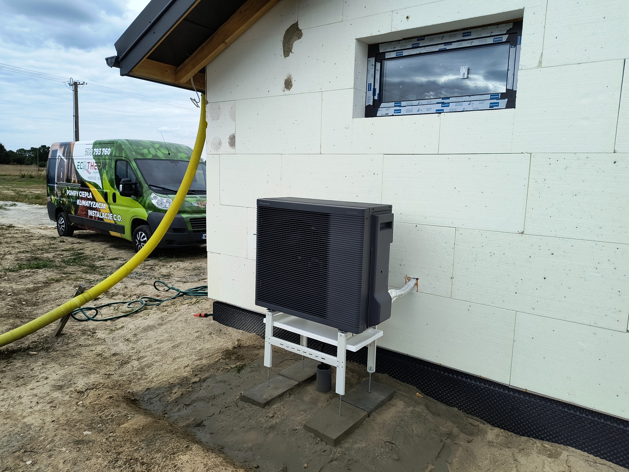 Zamontowana pompa ciepła Panasonic Aquarea na elewacji budynku z izolacją styropianową. Żółty wąż obok, w tle firmowy bus EcoTherm.