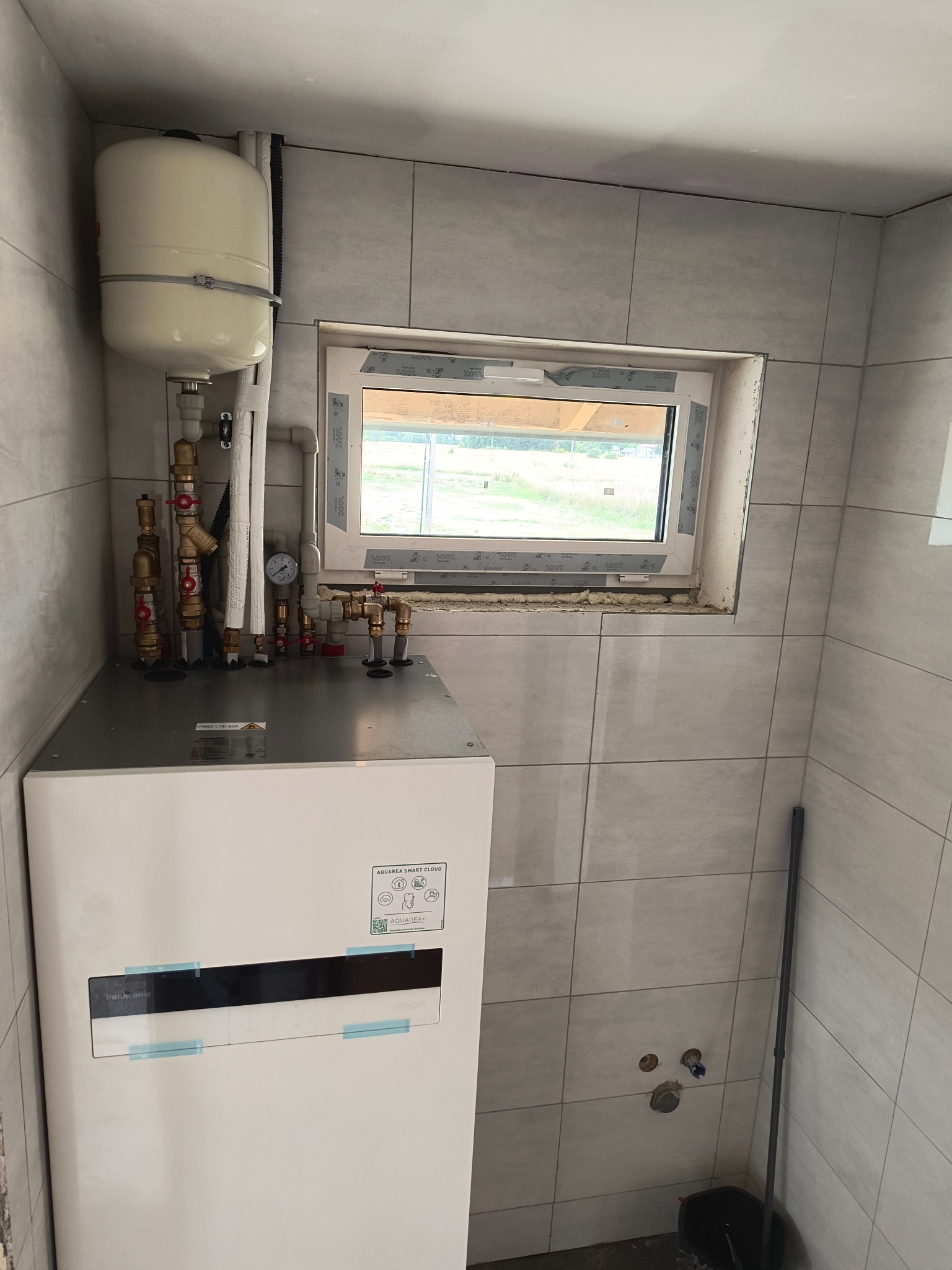 Biała pompa ciepła Panasonic Aquarea z zbiornikiem wyrównawczym, zamontowana w pomieszczeniu z szarymi płytkami, widok z przodu, instalacja w trakcie wykańczania.