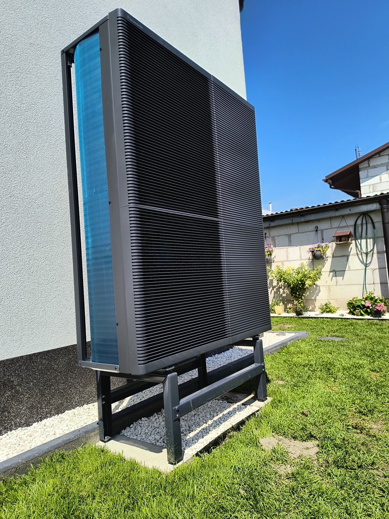 Zewnętrzna jednostka pompy ciepła Panasonic na posesji, ustawiona na ramie. Widoczne elementy instalacji, trawnik i fragment budynku w tle. Słoneczny dzień.