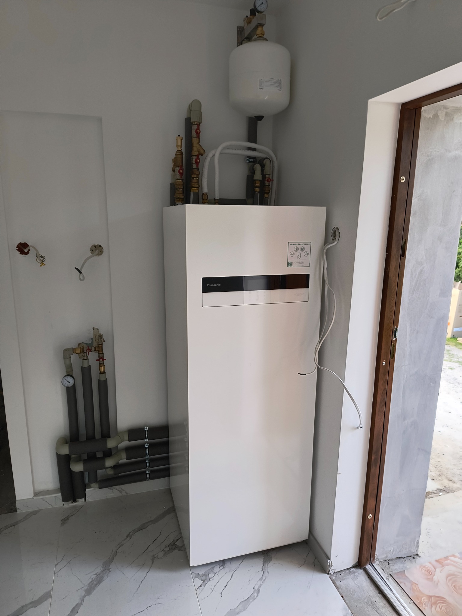 Biała pompa ciepła Panasonic Aquarea w pomieszczeniu z marmurową podłogą, widoczne rury i manometry. Częściowo widoczne drzwi z drewnianą ościeżnicą.