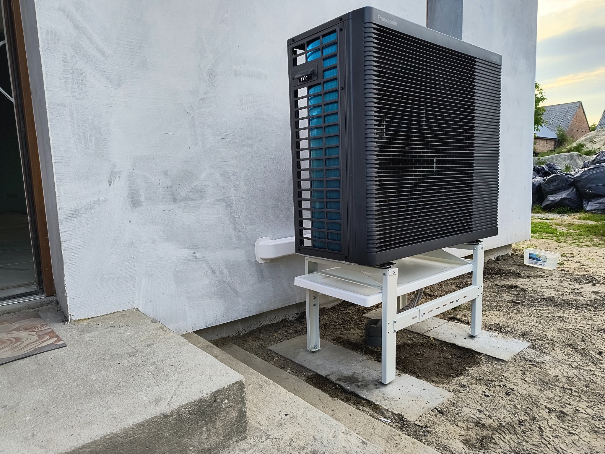 Czarna pompa ciepła Panasonic zamontowana na białym stelażu przy białej ścianie budynku. Widoczne elementy instalacji i otoczenie posesji w trakcie prac wykończeniowych.