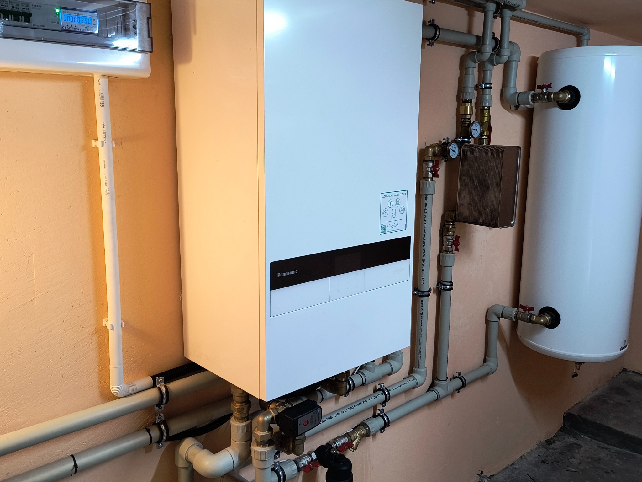 Biały piec Panasonic Aquarea Smart Cloud z szarymi rurami instalacji grzewczej na beżowej ścianie. Widoczny manometr i zbiornik. Elementy instalacji, kotłownia.