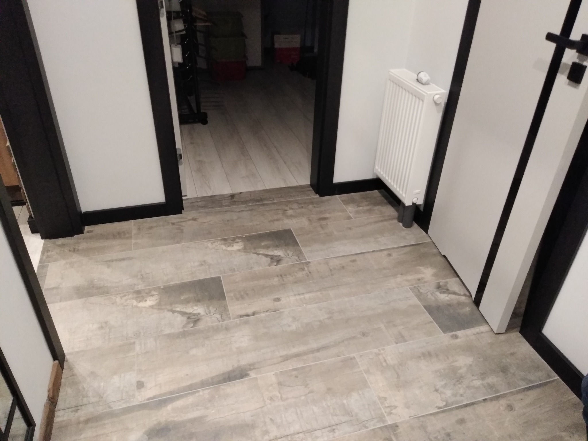 Ułożona podłoga z płytek imitujących drewno, biały grzejnik i czarne obramowania drzwi w jasnym wnętrzu.