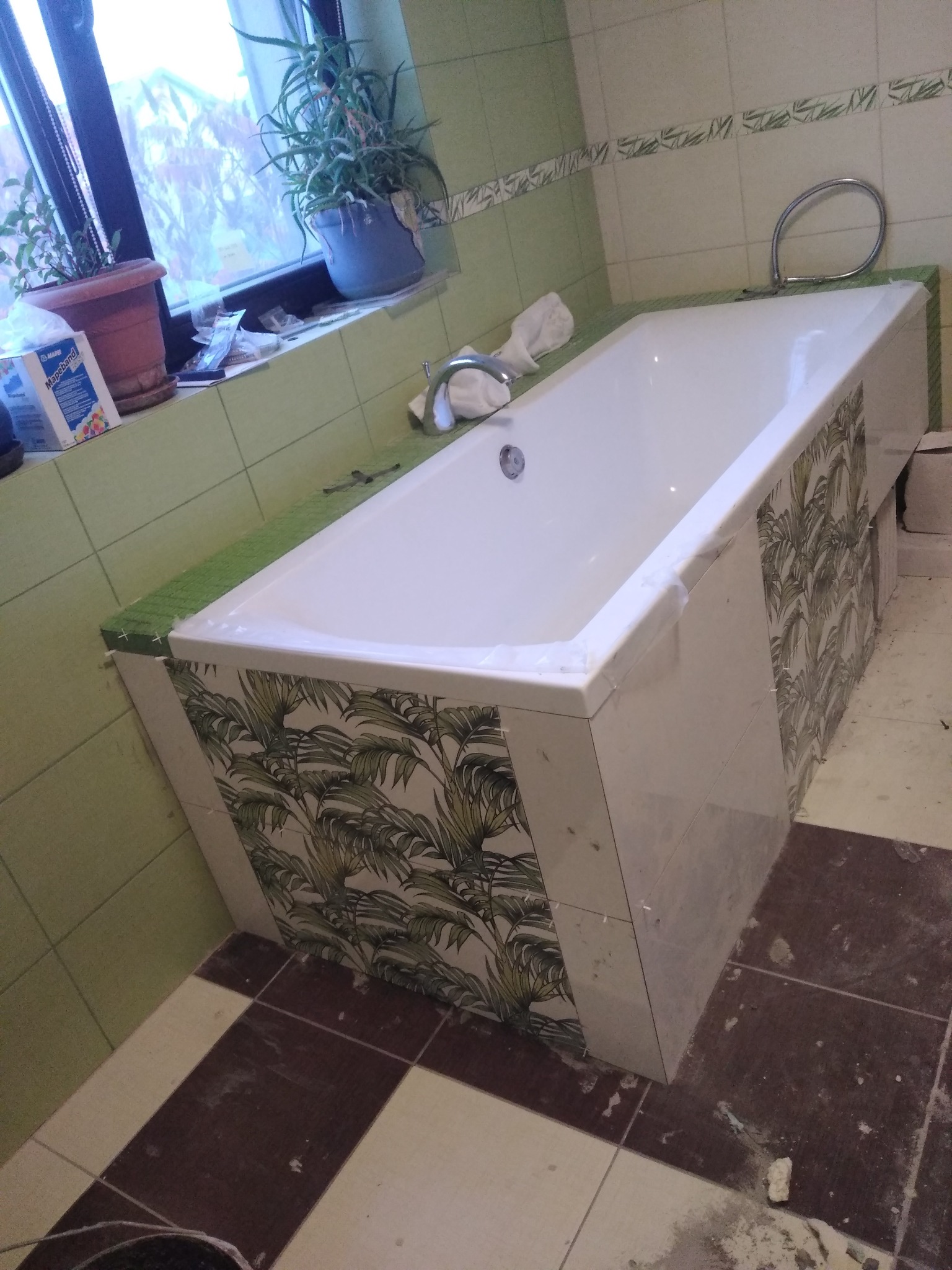 Biała wanna obudowana płytkami z motywem liści palmowych, fragment łazienki w trakcie remontu, widoczne rośliny doniczkowe na parapecie okna.