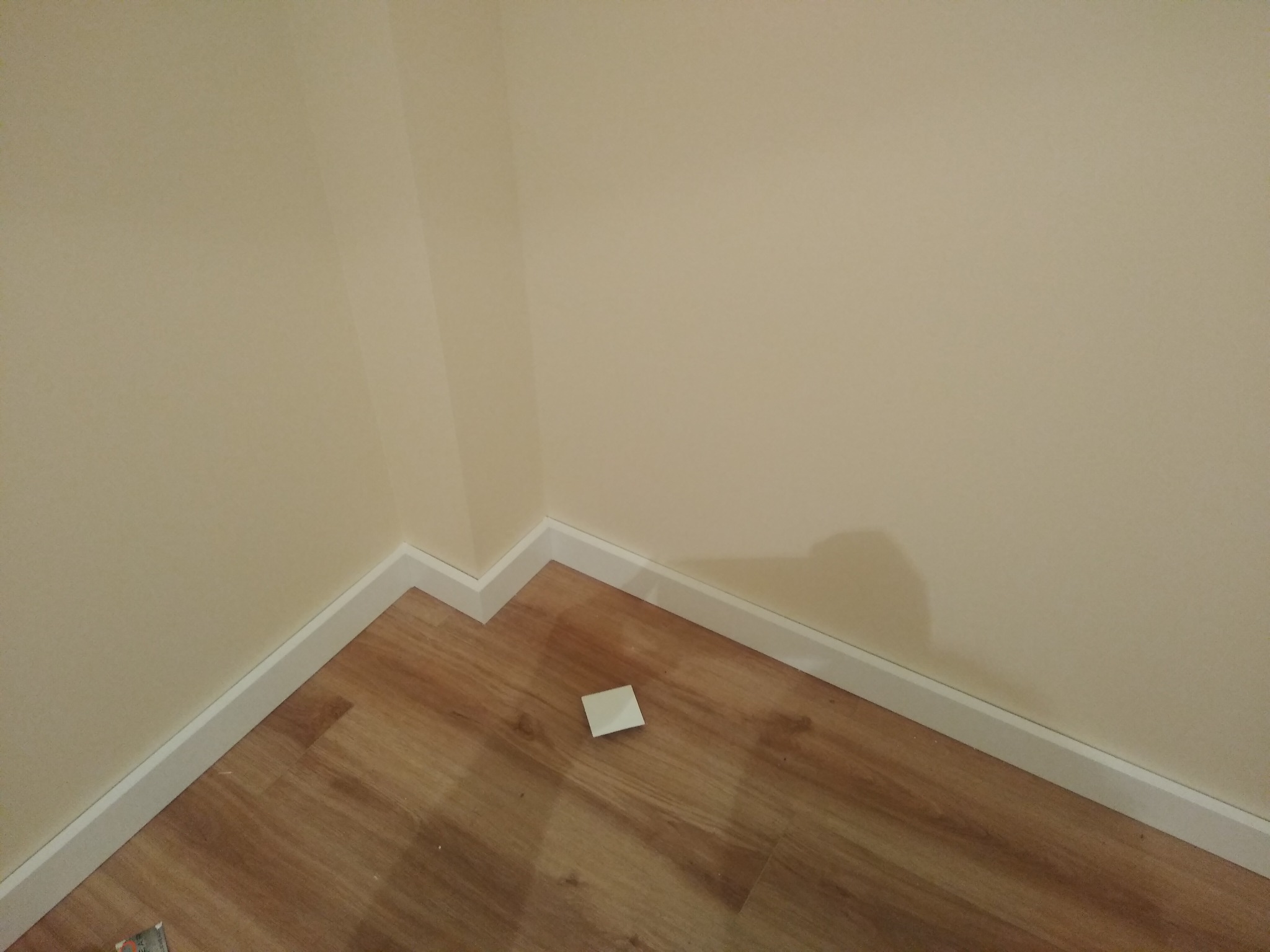 Fragment pokoju po remoncie: drewnopodobna podłoga z białymi listwami przypodłogowymi przy kremowej ścianie; na podłodze leży mały kwadratowy element.