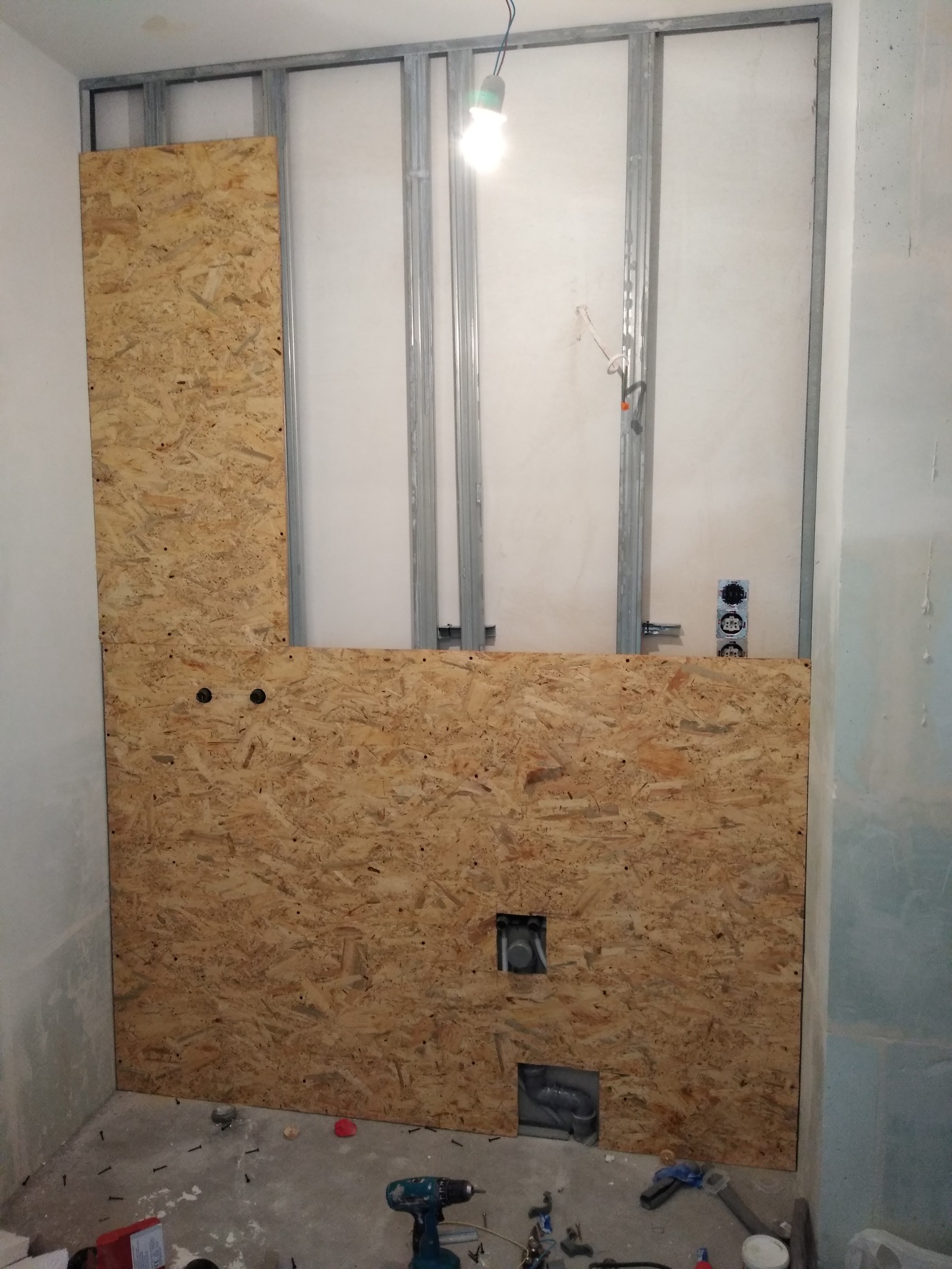 Wewnętrzna ściana z płyt OSB zamontowana na metalowym stelażu, z wyciętymi otworami na instalacje, widoczne narzędzia i śruby na podłodze.