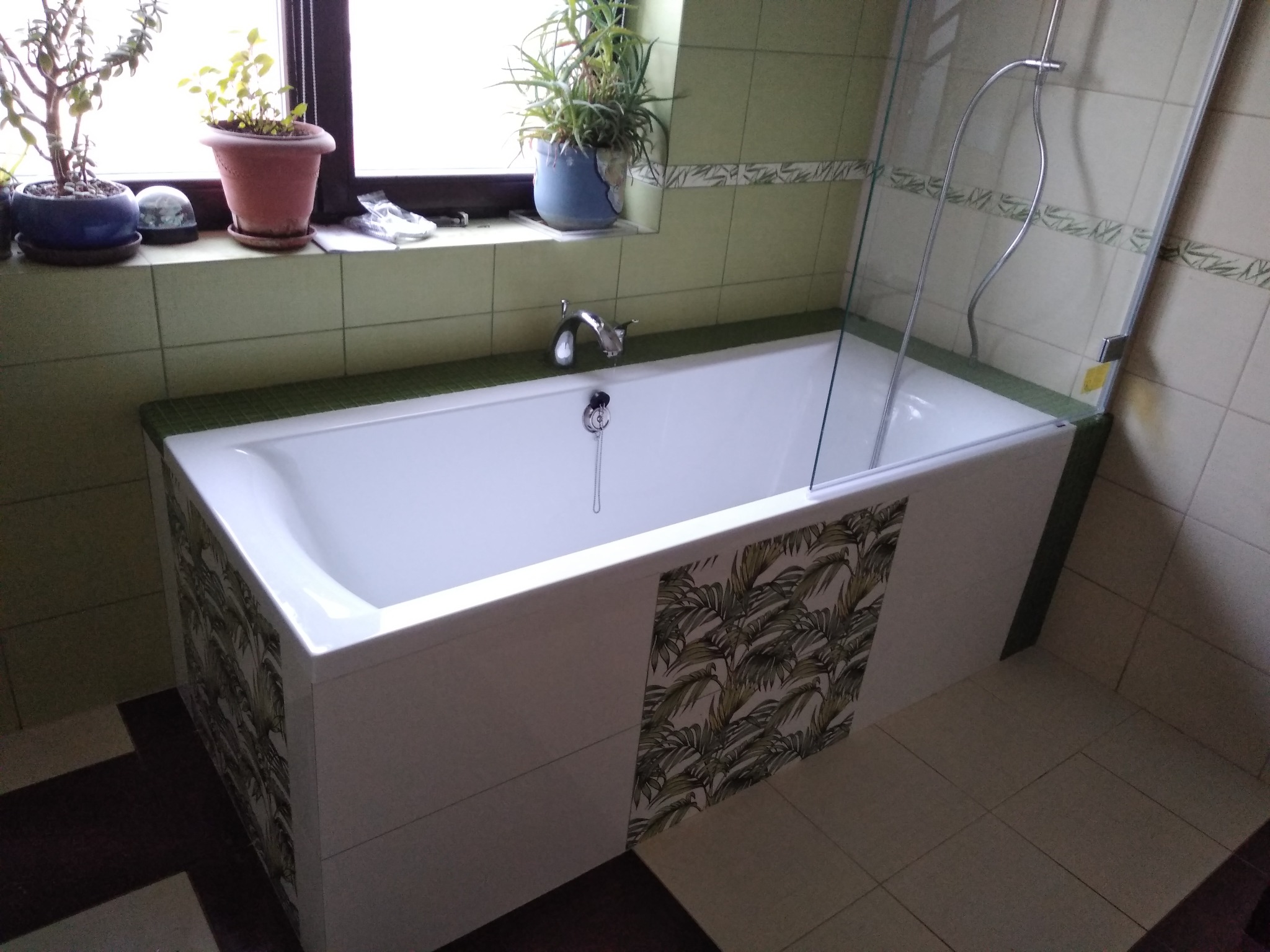 Nowoczesna wanna z parawanem nawannowym, obudowana płytkami z motywem tropikalnych liści, uzupełniona armaturą i roślinami doniczkowymi na parapecie okiennym.