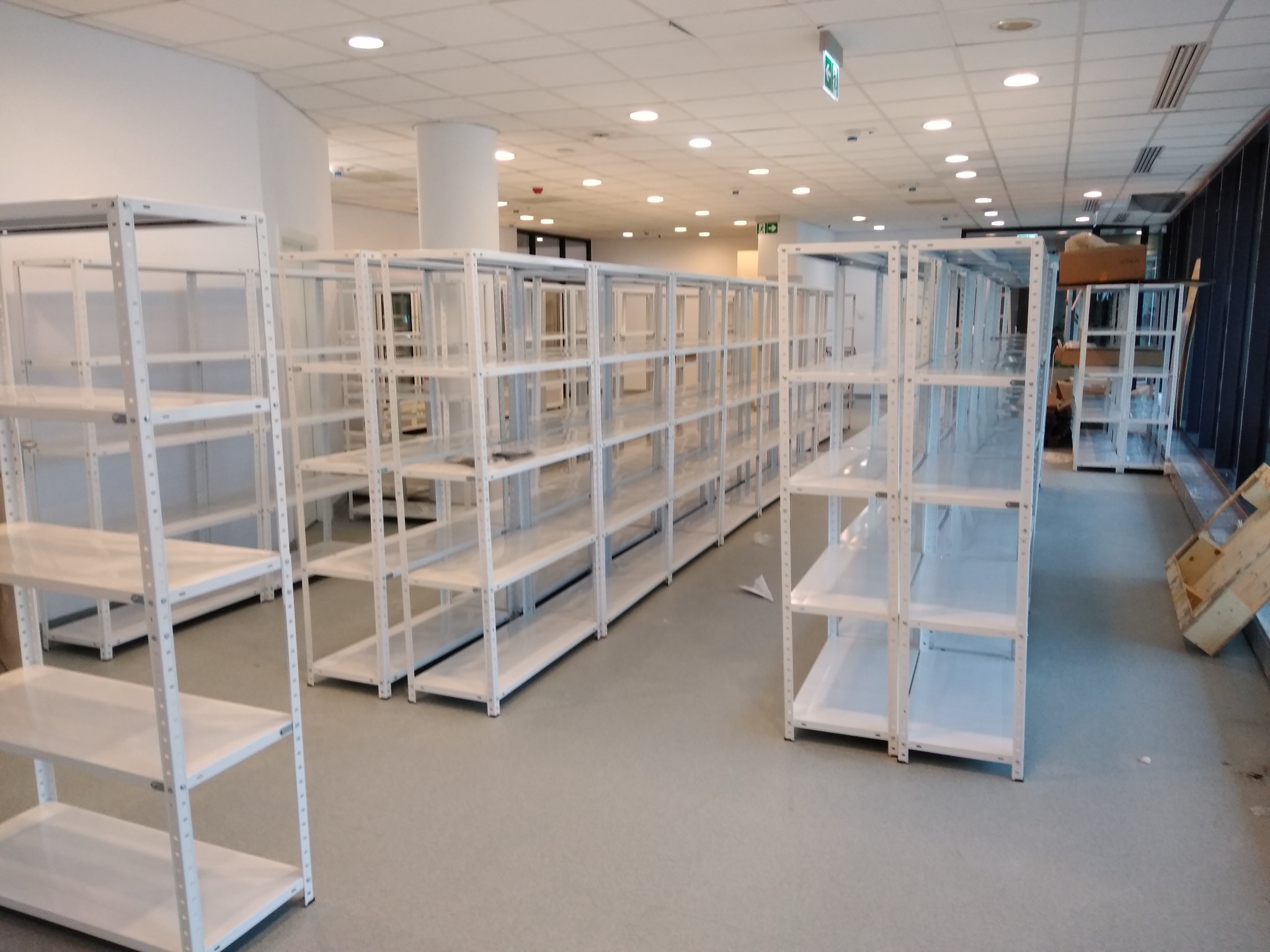 Długi rząd białych, metalowych regałów magazynowych z pustymi półkami, ustawionych w jasnym, przestronnym pomieszczeniu z kolumnami i podwieszanym sufitem, w tle widoczne kartony i drewniana skrzynka.