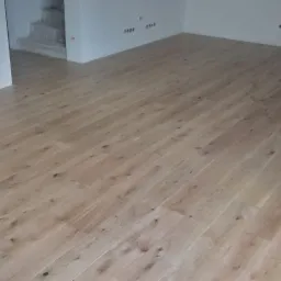 Jasne, drewniane panele podłogowe ułożone w nowym pomieszczeniu, widoczne fragmenty ściany i schodów w tle.