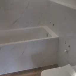 Wykończona łazienka z wanną w zabudowie, wiszącą toaletą i białymi płytkami imitującymi marmur na ścianach oraz panelami podłogowymi przypominającymi drewno.