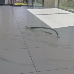 Białe, marmurowe płytki podłogowe ułożone w dużym, jasnym pomieszczeniu z widocznym wężem instalacyjnym wystającym z otworu w podłodze, w tle przesuwne drzwi tarasowe i zieleń.