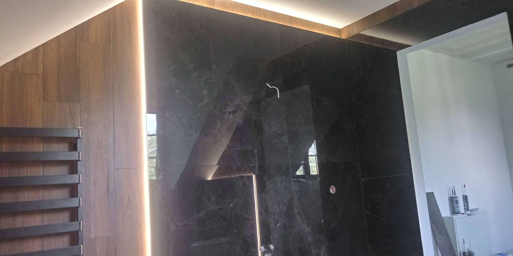 Nowoczesna łazienka z czarnymi marmurowymi płytkami, drewnopodobnymi panelami, podświetleniem LED i grzejnikiem drabinkowym.