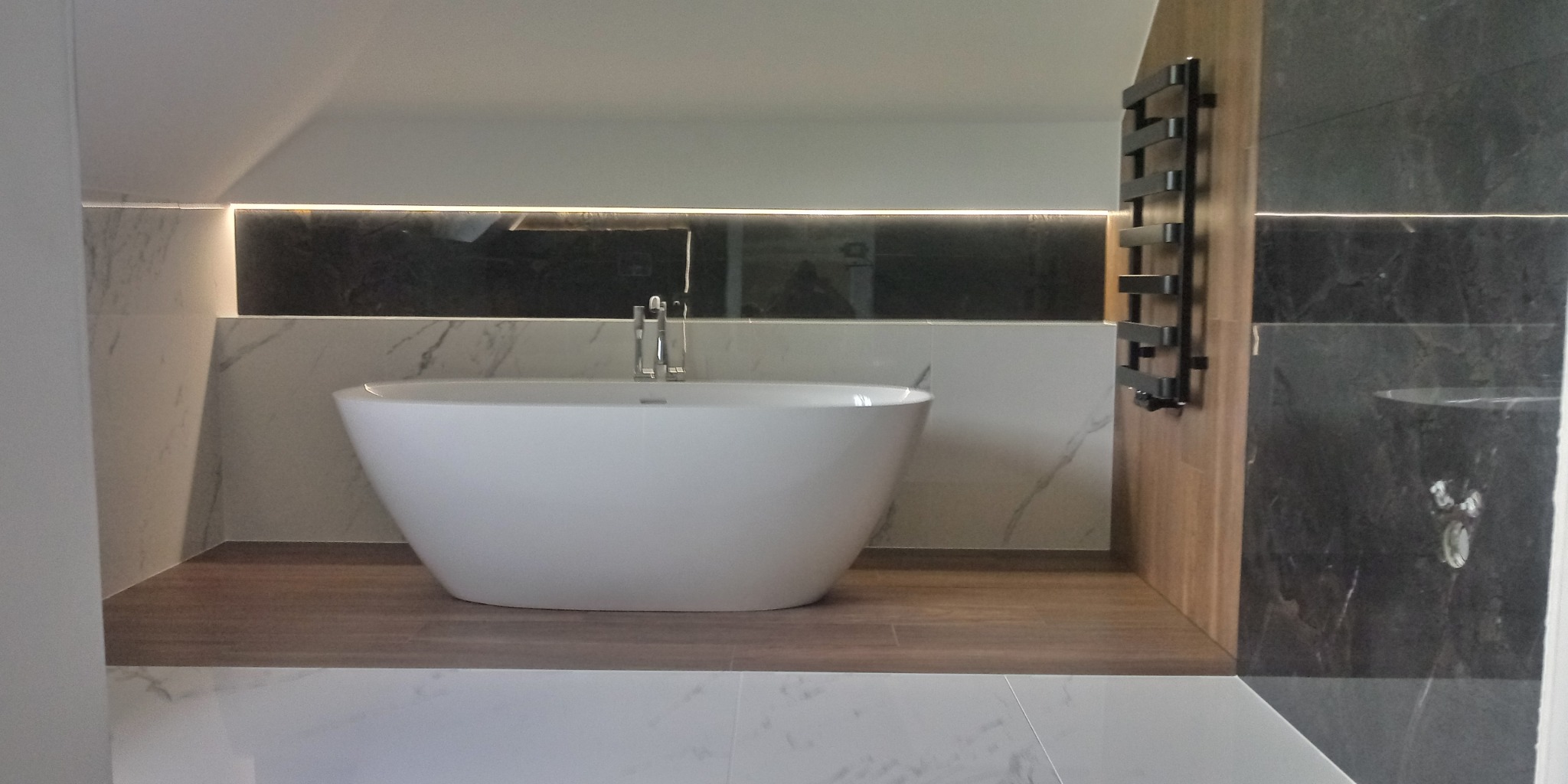 Wolnostojąca wanna owalna z chromowaną baterią, podświetlona wnęka z czarną płytką, grzejnik drabinkowy w kolorze czarnym, marmurowe płytki na ścianie i podłodze, drewnopodobny podest.