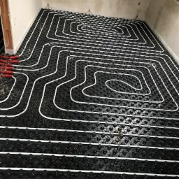 Białe rurki ułożone w pętle na czarnej macie podłogowej z wypustkami, przygotowane do zalania wylewką pod ogrzewanie podłogowe, widoczne metalowe uchwyty mocujące rurki.