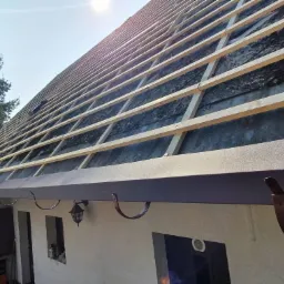 Dom w trakcie montażu łat dachowych i rynien w kolorze ciemnego brązu, widoczna drabina oparta o ścianę, zieleń w tle.