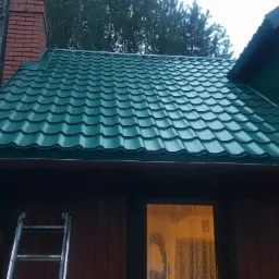 Dom z nowym, zielonym dachem z blachodachówki, brązowymi rynnami i ceglanym kominem, drabina oparta o ścianę.