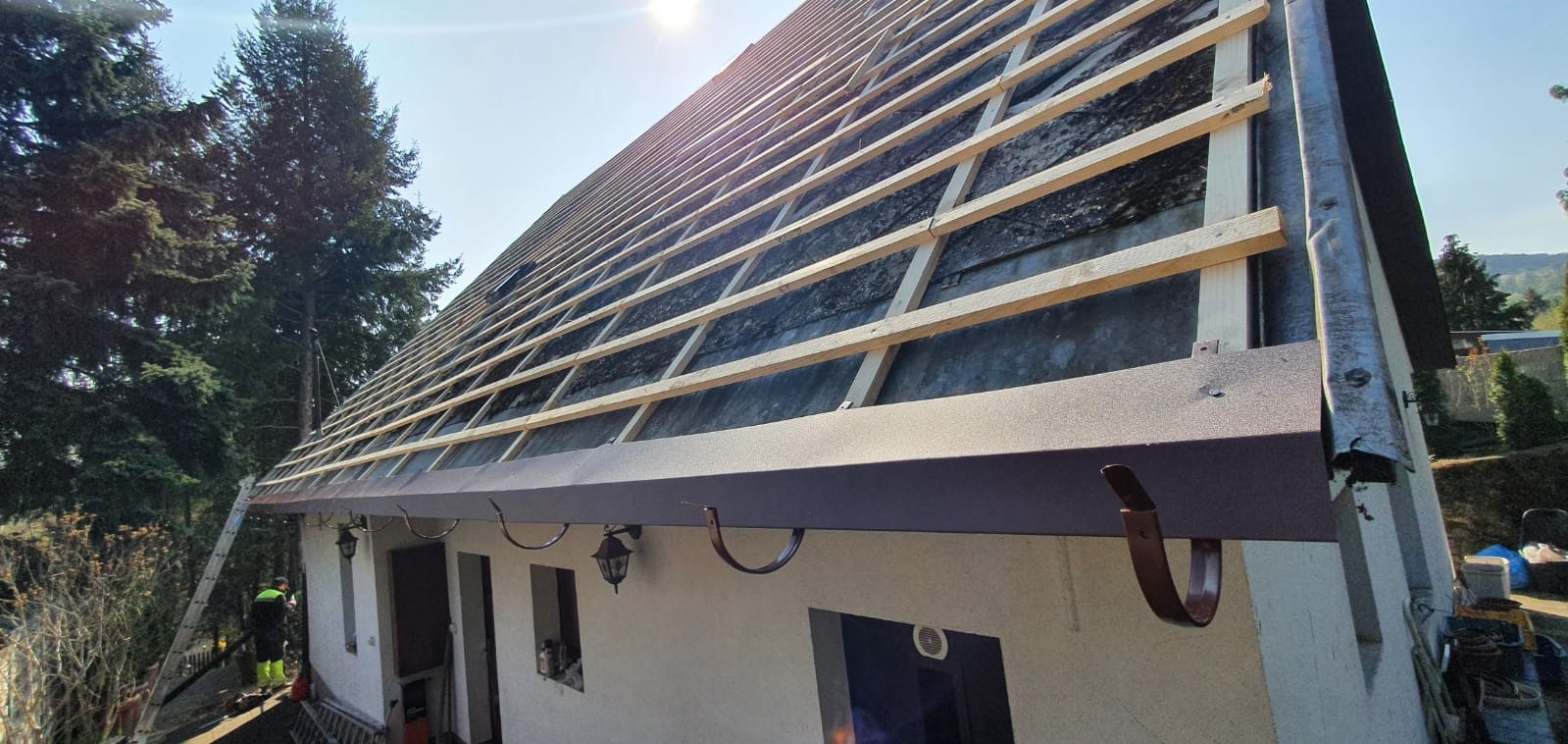 Dom w trakcie montażu łat dachowych i rynien w kolorze ciemnego brązu, widoczna drabina oparta o ścianę, zieleń w tle.