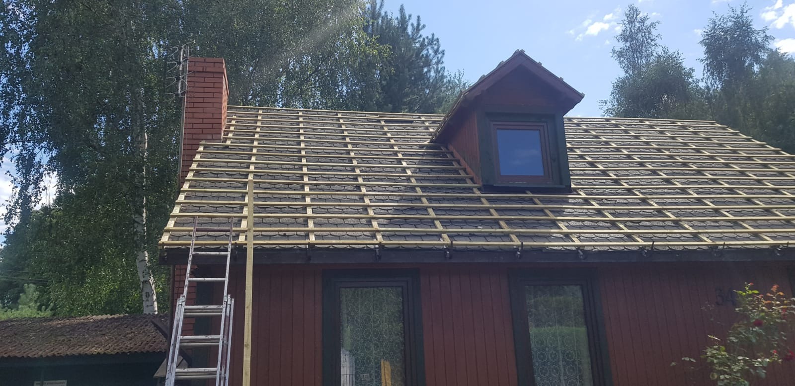 Dom z częściowo zdjętym starym pokryciem dachu, widoczne drewniane łaty pod nowe pokrycie, komin z czerwonej cegły i drabina oparta o ścianę.