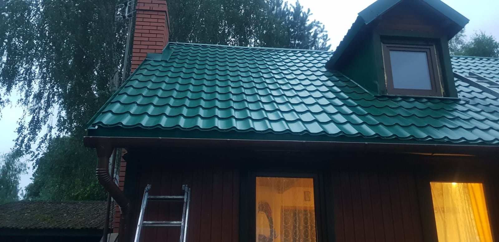 Dom z nowym, zielonym dachem z blachodachówki, brązowymi rynnami i ceglanym kominem, drabina oparta o ścianę.