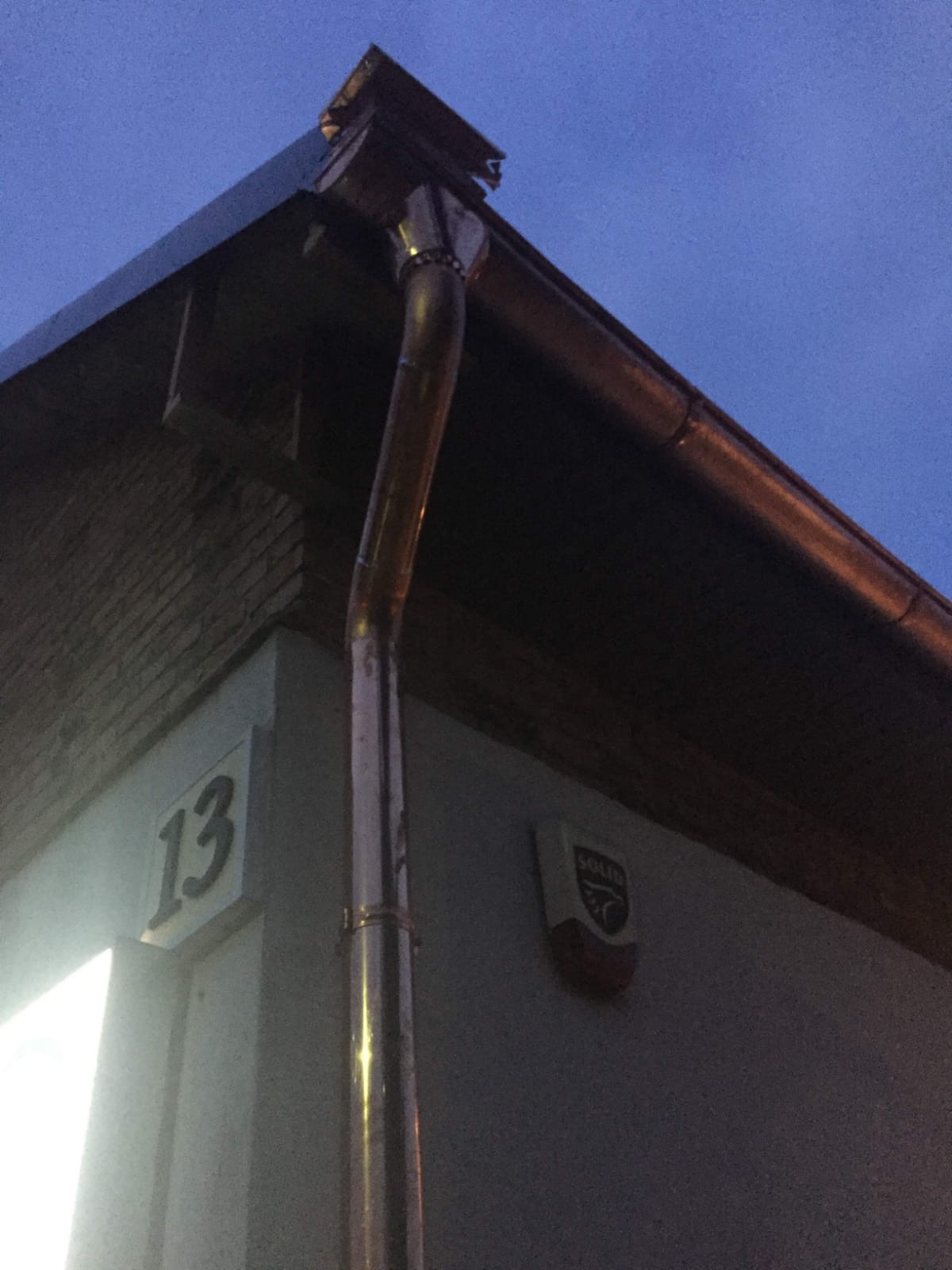 Miedziana rynna spustowa przy narożniku budynku z numerem 13, widoczny fragment dachu i alarm, nocne ujęcie.