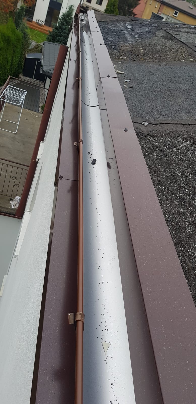 Widok z góry na dach z zamontowanym systemem rynnowym w kolorze brązowym i srebrnym, widoczne mocowania rynny spustowej i zabrudzenia na rynnie.