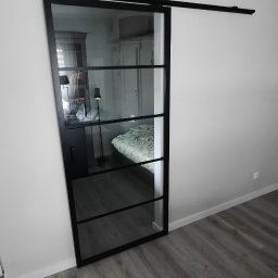 Usługi Remontowo-Budowlane Łukasz Purczyński - Szklane drzwi przesuwne w czarnej ramie, zamontowane na szynie, oddzielające pokój z widocznym łóżkiem i szafką nocną.