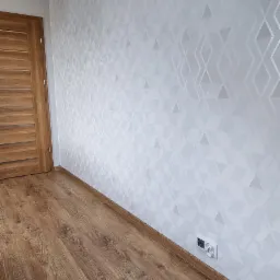 Wnętrze pokoju po remoncie: drewniana podłoga, ściana pokryta białą, geometryczną tapetą z efektem 3D, dębowa futryna drzwiowa.